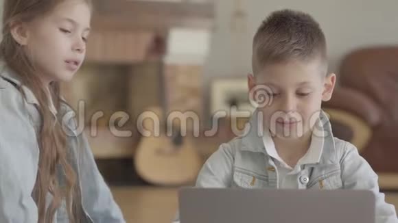 肖像可爱的专心致志的男孩正在电脑上打字或玩耍他的双胞胎妹妹饶有兴趣地看着什么视频的预览图