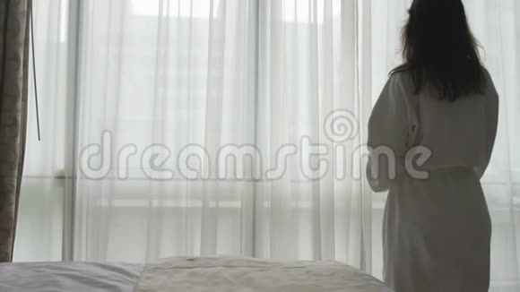 穿着浴袍的年轻女子揭开窗帘张开双臂从窗户望去视频的预览图