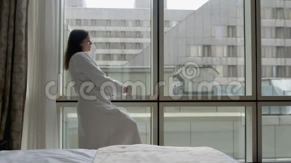 穿着白色浴袍的年轻女子站在酒店公寓的窗户旁手里拿着智能手机和一杯咖啡视频的预览图