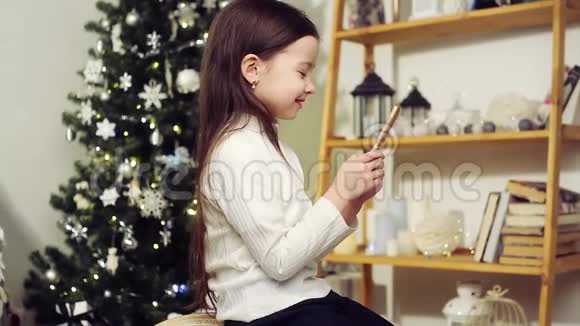 可爱的女孩在圣诞树的背景上带着电话视频的预览图