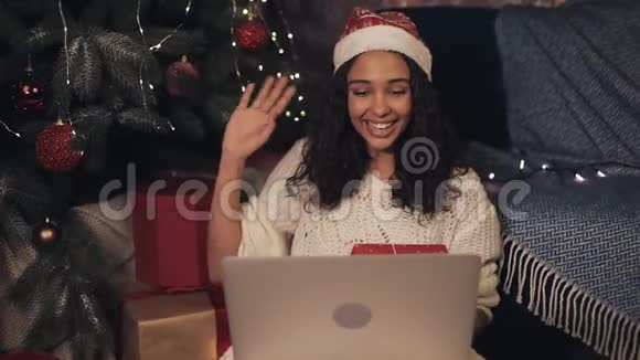 迷人的年轻微笑女孩戴着桑塔斯帽子手提电脑上有视频通话坐在家附近的圣诞树妇女视频的预览图