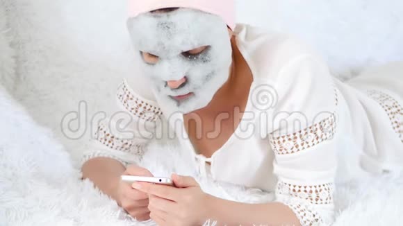 一个躺在沙发上的年轻女人用她的智能手机上网女人用泡泡面膜敷脸皮视频的预览图