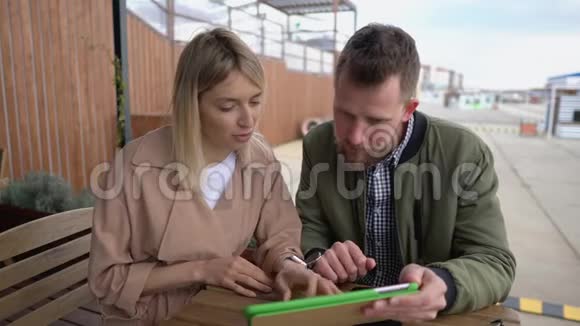 年轻夫妇正坐在户外的咖啡馆里通过平板电脑浏览互联网网站视频的预览图