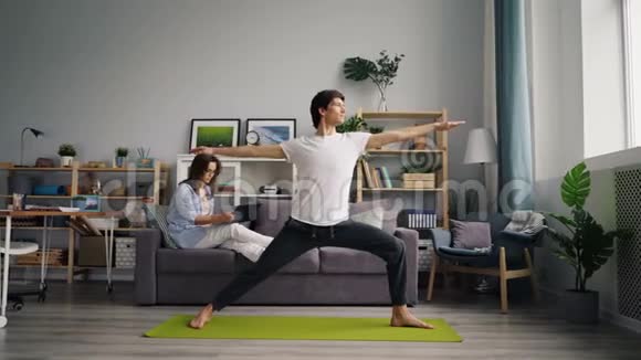 混合种族的年轻人在家里锻炼当妻子在电话里聊天时做瑜伽视频的预览图