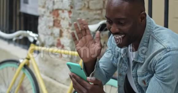 一个快乐的非裔美国人坐在时髦的自行车旁时通过他的智能手机电话挥挥手微笑视频的预览图