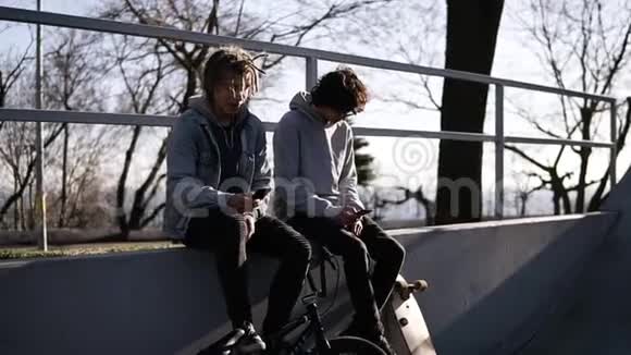 两个朋友滑板车和bmx骑手正坐在城市滑板公园的高栏杆上使用他们的滑板车视频的预览图