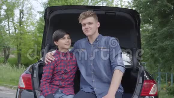 肖像年轻的父亲和他的儿子坐在户外的汽车后面拥抱男孩的男人看着每个人视频的预览图