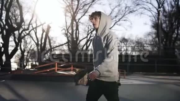穿着灰色连帽衫的年轻滑板手拿着滑板在滑板公园里散步侧视慢动作视频的预览图