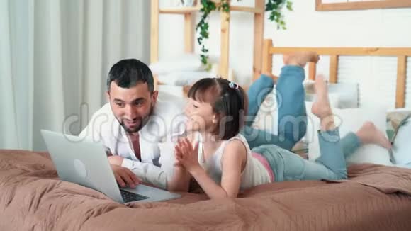 爸爸和可爱的孩子在家里用笔记本电脑看有趣的视频躺在床上慢动作视频的预览图