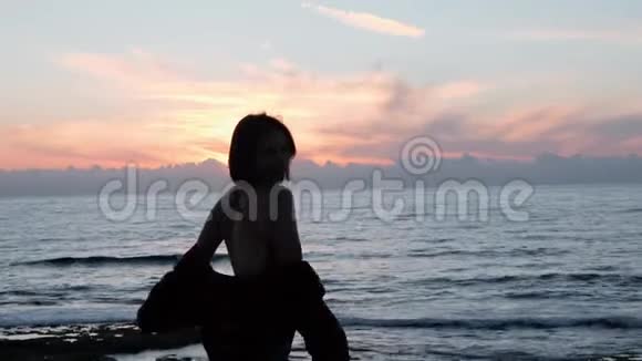 太阳落山时为照相机摆姿势的棕发辣妹年轻女子的剪影微笑着在岩石海岸旋转吸引力强视频的预览图