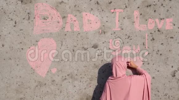 一个穿着粉红色头巾的女孩用粉笔在墙上写字爸爸我爱你视频的预览图