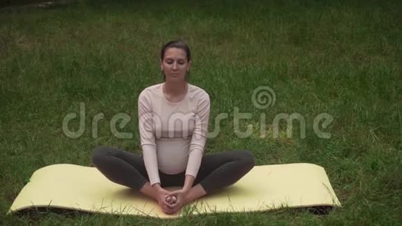 一位孕妇在公园里的地毯上练习瑜伽做ASDBADDAKonasana蝴蝶姿势视频的预览图