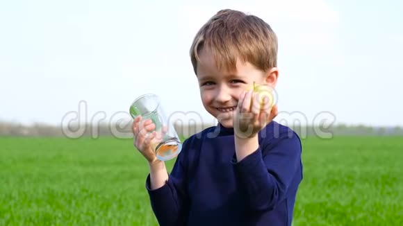 孩子拿着一个绿色的苹果和一杯苹果汁男孩在大自然的背景下喝果汁笑视频的预览图
