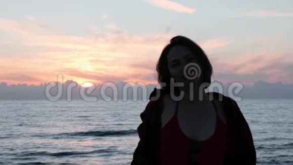 年轻迷人的女孩穿着红色上衣和牛仔裤透过相机在海岸看炎热的女人在黄昏时眺望远方年轻女孩视频的预览图