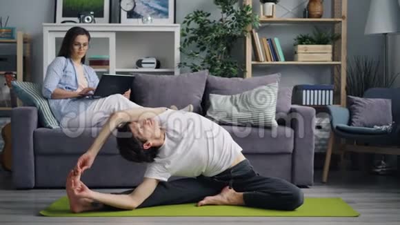 灵活的年轻人做瑜伽而女友则坐在沙发上用笔记本电脑视频的预览图