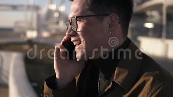 戴着时髦眼镜和耳环的开朗微笑的男人正用他的手机说话坐在靠近栏杆的外面视频的预览图