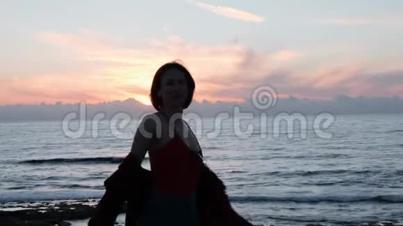 太阳落山时为照相机摆姿势的棕发辣妹年轻女子的剪影微笑着在岩石海岸旋转吸引力强视频的预览图