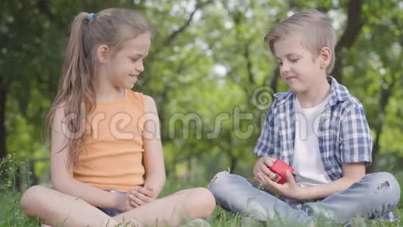 漂亮的小女孩和坐在草地上的英俊男孩在一起男孩拿着一个红色的小盒子打开它视频的预览图