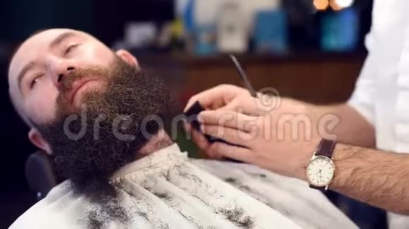 重点是用电动剃须刀切割胡须用理发师的手和梳理留着卷曲黑胡子的人的正面景色视频的预览图