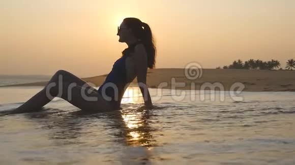 年轻女子躺在沙滩上被海浪冲走日落时分可爱的女孩在热带海岸放松视频的预览图