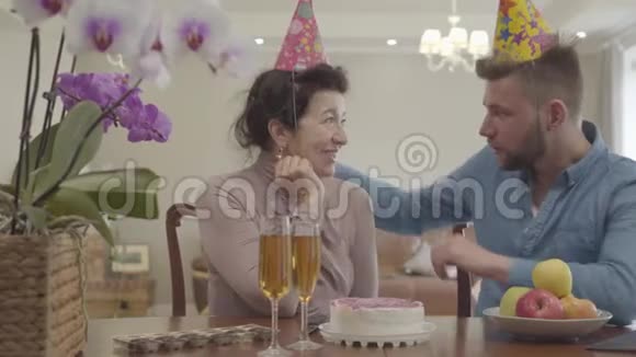可爱的奶奶和成年孙子坐在桌子旁头上戴着生日帽切蛋糕的男人女人视频的预览图