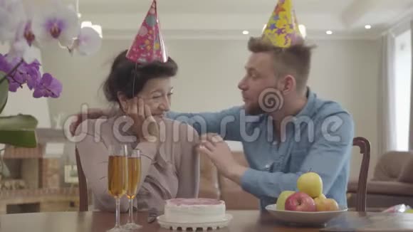 肖像成熟的女人和成年的孙子坐在桌子旁头上戴着生日帽在桌子上视频的预览图