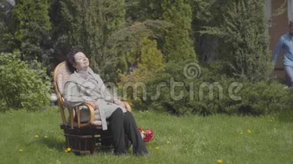 肖像可爱成熟的女士坐在摇椅上享受阳光成年孙子带着水来视频的预览图