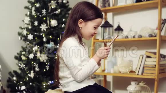 可爱的女孩在圣诞树的背景上带着电话视频的预览图