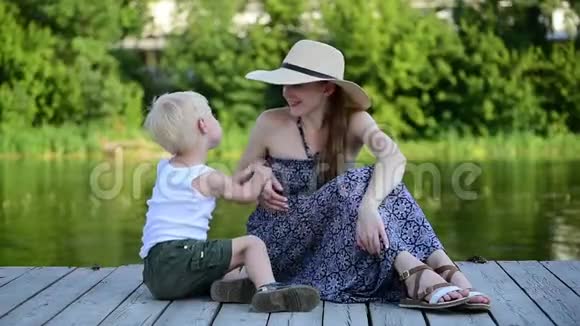 妈妈和她的金发儿子在河边的码头上玩和聊天夏日视频的预览图