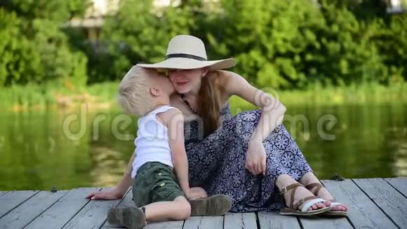 妈妈和她金发的儿子在河边的码头上拥抱和聊天夏日视频的预览图