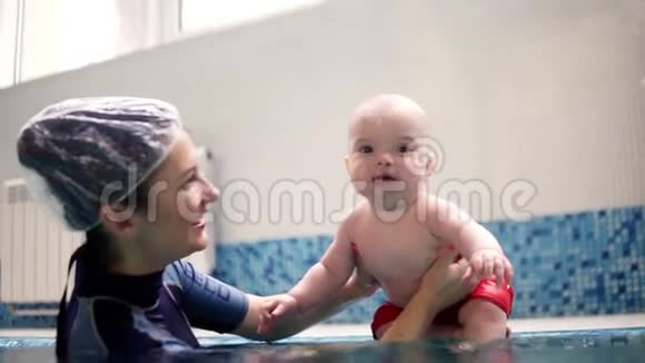 一个穿红色短裤的可爱的托德勒坐在游泳池的一侧然后在他的母亲或视频的预览图