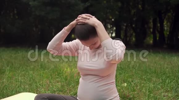 孕妇在公园里地毯上做瑜伽坐在莲花的位置热身脖子和肩膀视频的预览图