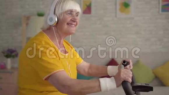 戴耳机的活跃老年妇女在运动自行车上进行有氧训练视频的预览图