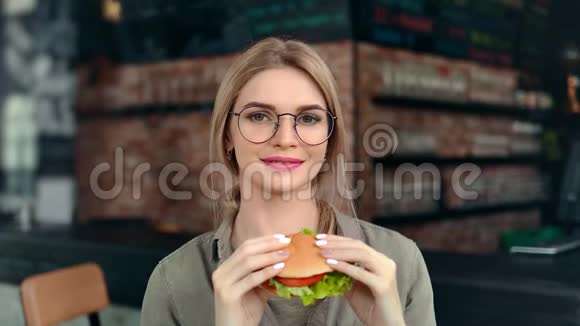 戴着眼镜可爱微笑的年轻女子的肖像咬着开胃的新鲜汉堡中等特写镜头视频的预览图