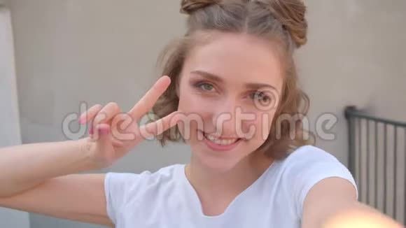 一位年轻可爱的白种人女性的特写镜头她的头发包在手机上自拍露出两只手指看着视频的预览图