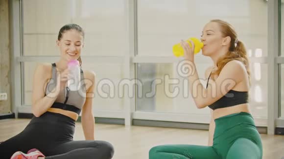 两个穿着运动服的微笑女人坐在健身房里从瓶子里喝水说话视频的预览图