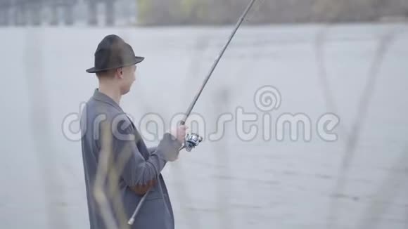 清晨年轻的渔夫穿着一件夹克头上戴着一顶帽檐在河边钓着一条旋转着的蜘蛛鱼视频的预览图