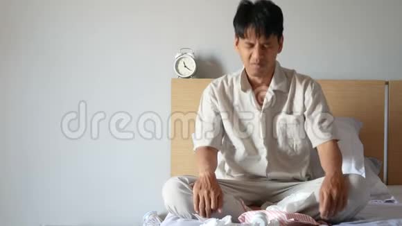 4K穿着睡衣的亚洲男人因过敏而吸鼻涕用纸巾擦拭粘液坐在卧室的床上视频的预览图