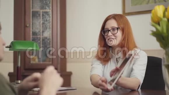 戴眼镜的漂亮红发年轻女心理学家正在咨询她的病人并在平板电脑上显示信息视频的预览图
