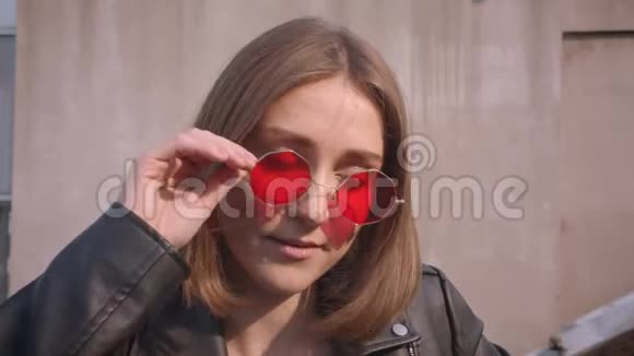 年轻可爱的白种人女孩穿着皮夹克戴着红色太阳镜开心地摆在面前的特写照片视频的预览图