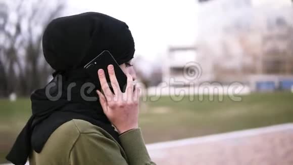 一位年轻的穆斯林妇女戴着传统的头巾在公园的智能手机上和朋友聊天一位女士走在路上视频的预览图