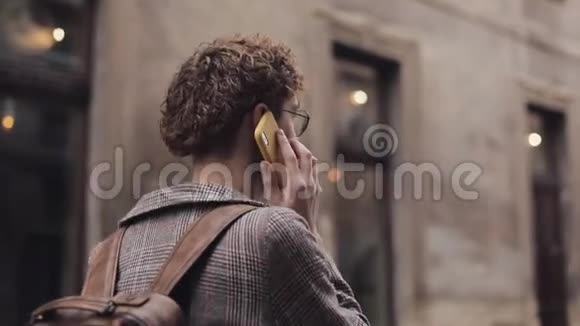 戴眼镜的年轻人侧后景带包在电话边走在老城街通信视频的预览图