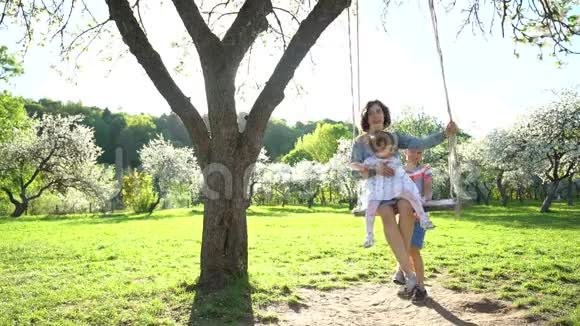 儿子在盛开的花园里荡着母亲和妹妹的木秋千静态射击视频的预览图