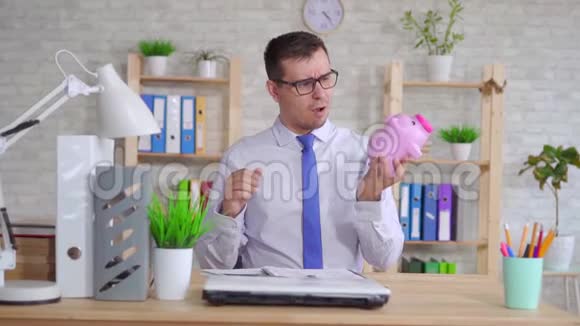 穿着衬衫的男人坐在办公室的笔记本电脑旁手里拿着一个储蓄罐视频的预览图