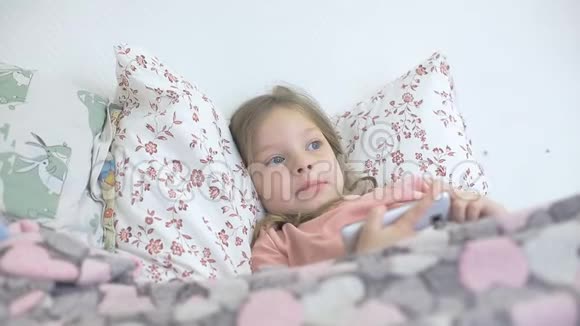 一个生病的小女孩和她的母亲躺在床上看着智能手机屏幕上的卡通片儿童和数字视频的预览图