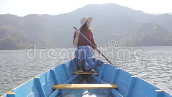 穿着红色衬衫的女孩戴着帽子戴着太阳镜坐在一艘蓝色的木船上手里拿着一只桨微笑着在湖边交谈视频的预览图
