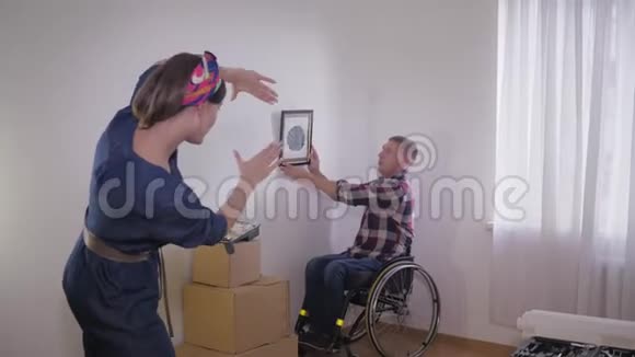 坐在轮椅上的有帮助的男人挂着照片而他的妻子用手指做框架并选择位置作为图像视频的预览图