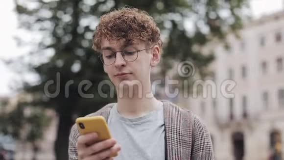 戴眼镜的年轻学生使用他的现代手机设备在屏幕上滚动微笑行走户外视频的预览图