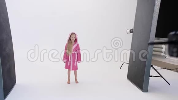 无法辨认的人拿着相机拍下兴奋的穿着浴衣的女孩在摄影棚白色背景上跳跃的照片视频的预览图