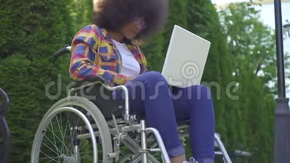 戴着非洲发型轮椅残疾的黑人女性戴着笔记本电脑在公园里晒太阳视频的预览图